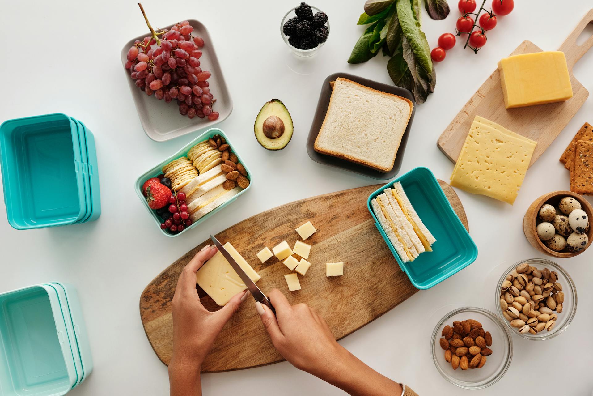 Person slicing cheese on a wooden board with various food items around on a white surface